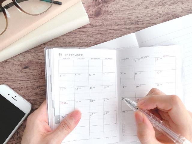 9月のカレンダーに予定を書き込んでいる手元、ペンと携帯電話、メガネがデスクの上に並んでいる