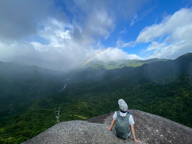 屋久島の岩山の上に座り雄大な山々と虹を眺めるトレッカー