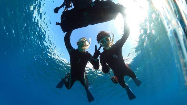 水中でスキューバダイビングをしている二人がピースサインを出し、明るい太陽の光が水面を通して差し込んでいるシーン