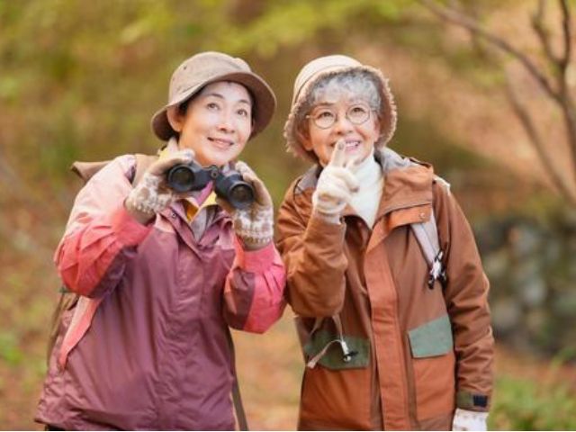 アウトドアウェアを着て、双眼鏡を手に森の中でバードウォッチングや自然観察を楽しむ笑顔のシニア女性2人。