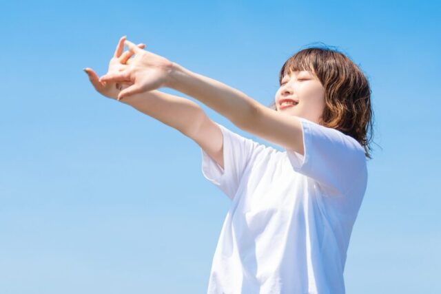 青空に向かって両腕を伸ばし笑顔の女性