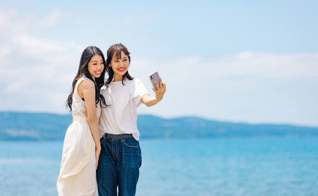 二人の女性がビーチでスマートフォンで自撮りをしている、青い空と海が背景に広がる風景
