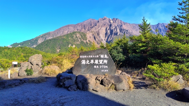 鹿児島の湯之平展望所にある石の標識と、背後に広がる山と青空の風景