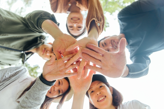 5人の人々が手を重ねてポーズを取っているシーン、カメラの下から撮影され、笑顔で協力の気持ちを示す