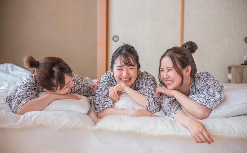 浴衣姿で民宿のベッドに寝転びながら笑顔で談笑する女性3人